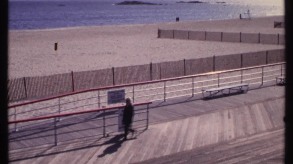Elevated 8mm view of a New London beachfront boardwalk as a distant pedestrian walks past signs and railings.
