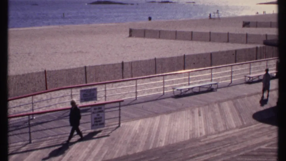 New London beachfront boardwalk with one pedestrian crossing foreground and another figure near an empty bench, with sand and water beyond.