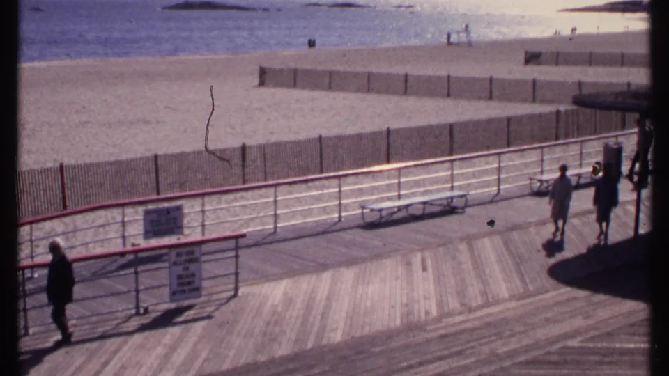 1969 New London shoreline boardwalk with multiple distant pedestrians, empty benches, railings, beach fencing, sand and ocean water.