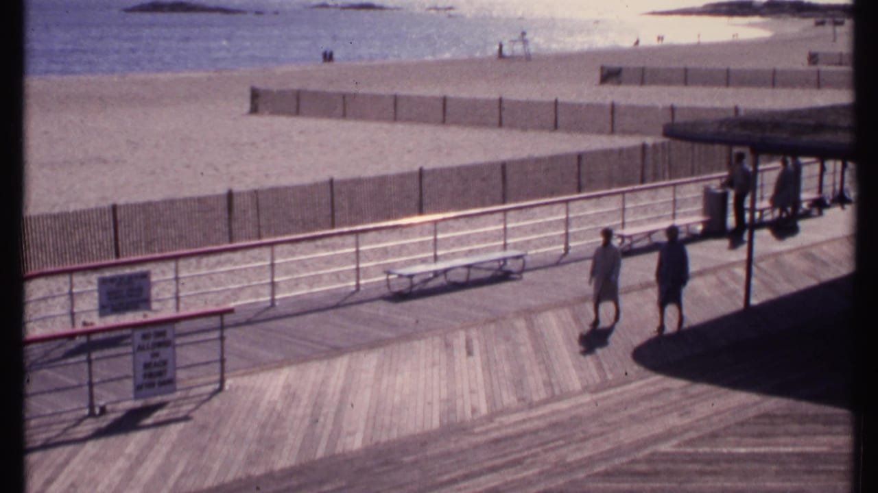 Beach boardwalk from above with walkers, benches, railing, fence, and sand beyond.