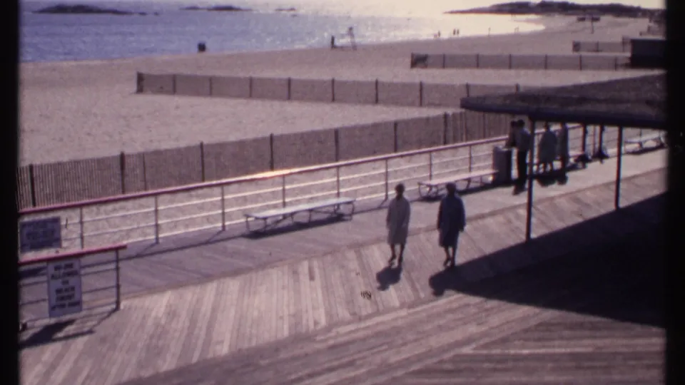 Elevated 1969 New London beach boardwalk view showing two adults walking toward a shaded pavilion area near benches and beach fencing.