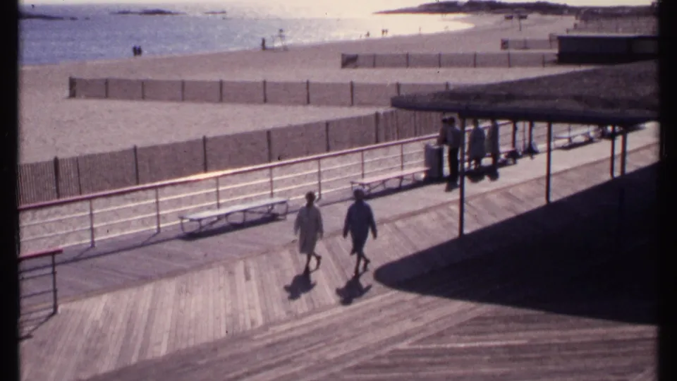 1969 New London seaside boardwalk with adults walking in light clothing, benches, railings, beach fencing, sand, and water beyond.