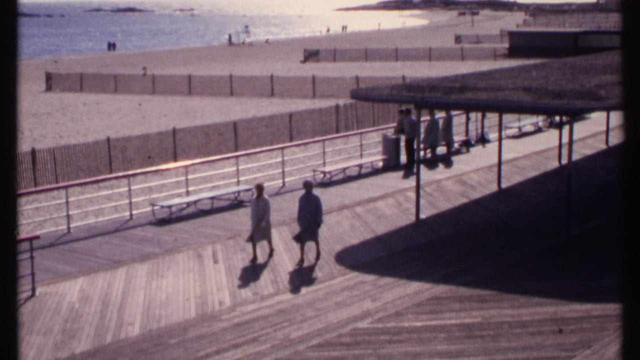 Two people walk across a sunlit boardwalk near a beach with railings, benches, and a covered structure.