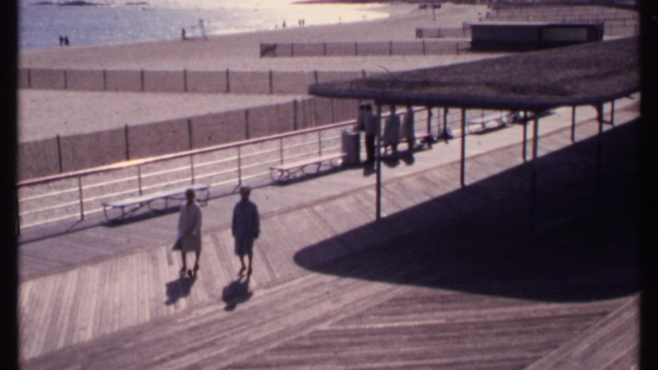 Two people walk slowly on the New London boardwalk in 1969, with benches, railings, beach sand, water, and distant figures visible.
