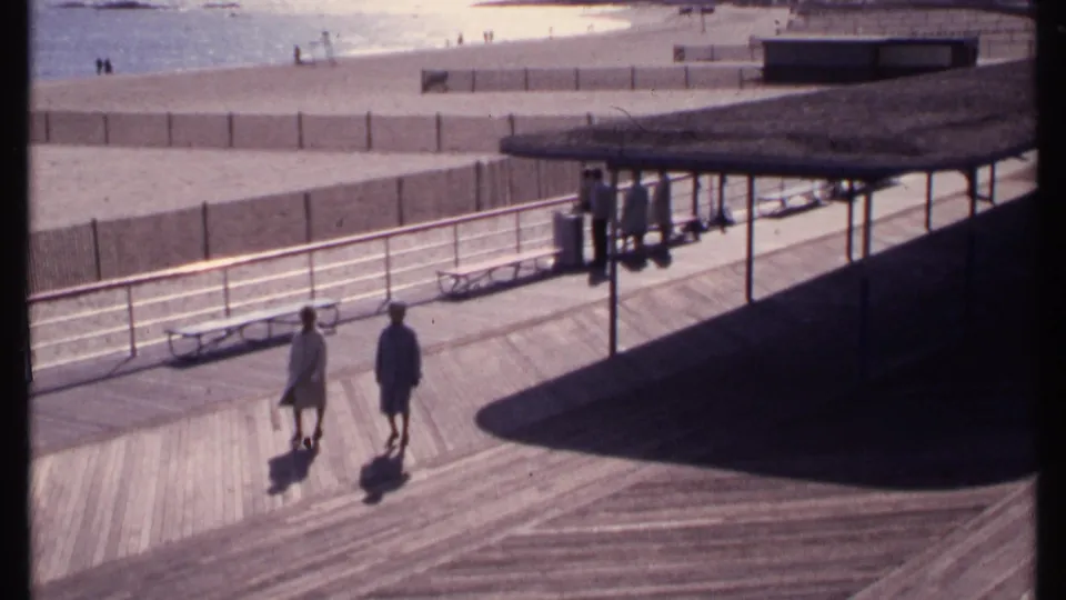 Two people walk slowly on the New London boardwalk in 1969, with benches, railings, beach sand, water, and distant figures visible.