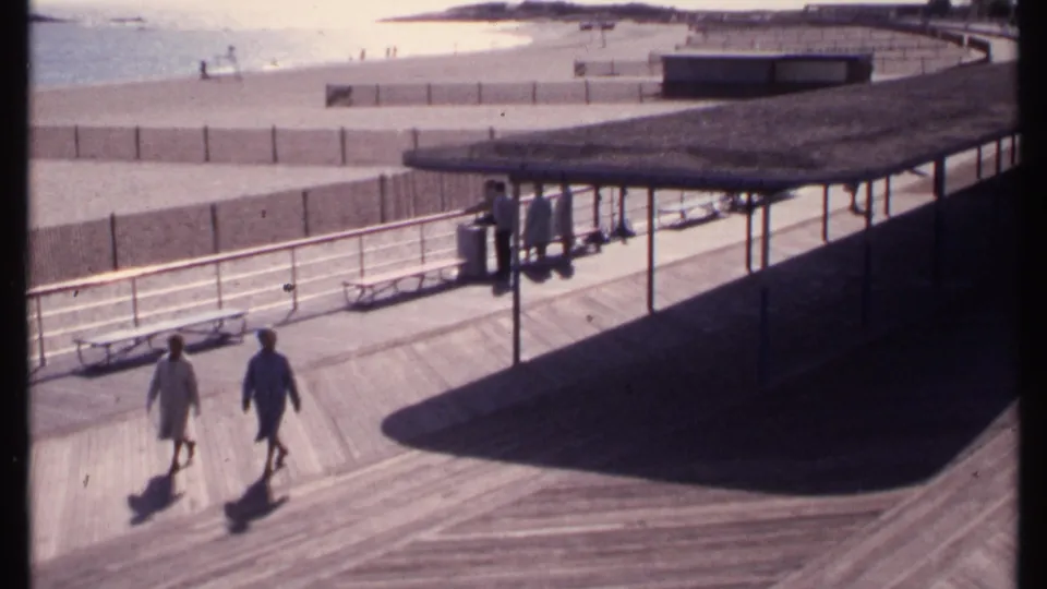 1969 boardwalk footage from New London shows two pedestrians passing below railings and benches beside the sandy beach.