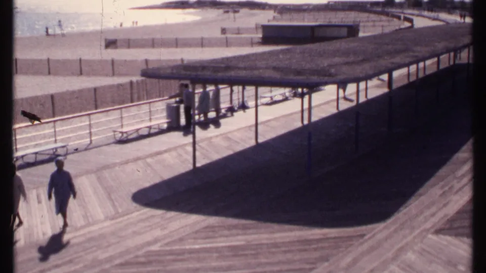 End of the 1969 New London boardwalk shot with one walker centered, another partly leaving frame, and beach structures behind them.