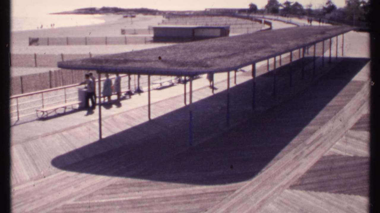 Covered seaside pavilion and boardwalk with beachgoers standing near railings and sand beyond.