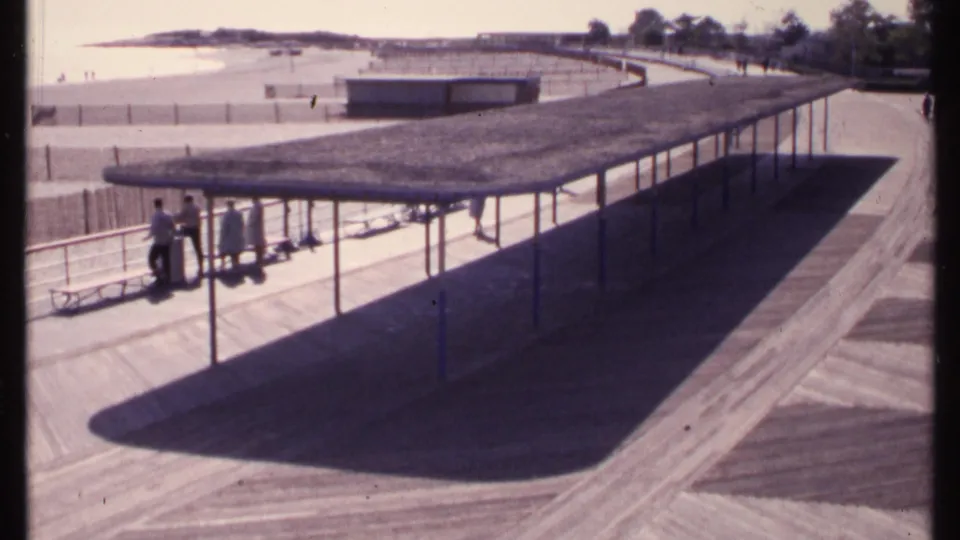 1969 New London shoreline frame showing the covered boardwalk pavilion, visitors in casual dress, benches, sandy beach, and distant water.