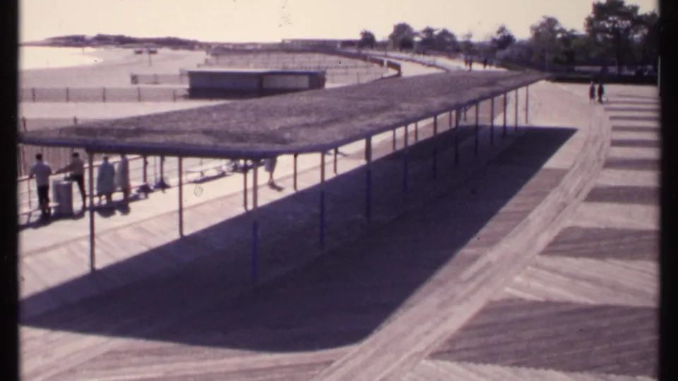1969 New London, Connecticut frame with a long pavilion shadow over the boardwalk, beachgoers at left, sand, shoreline water, and trees beyond.
