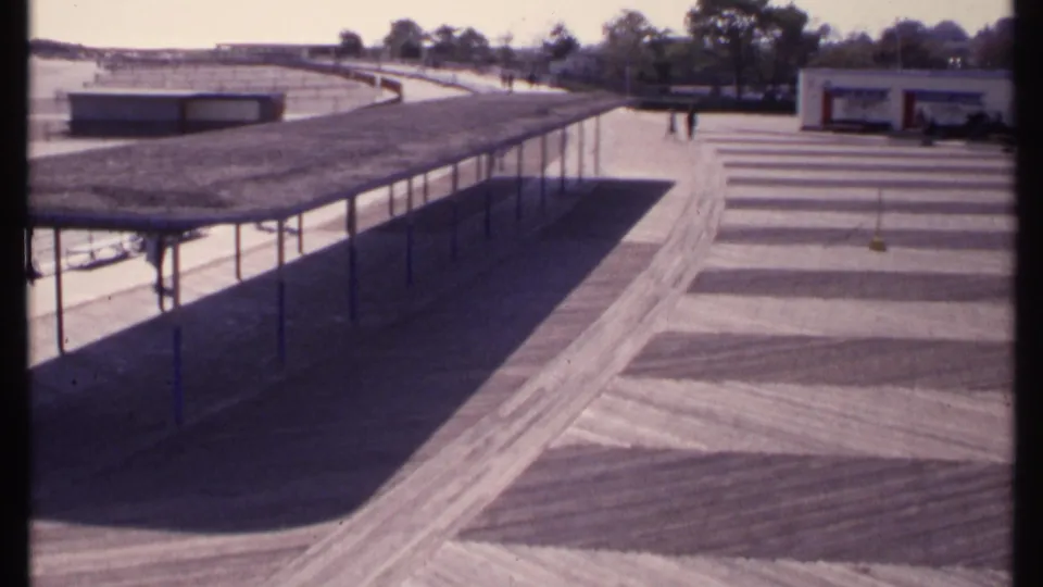 1969 home-movie view in New London showing a covered pedestrian shelter, open waterfront paths, low building, trees, and a few distant visitors.