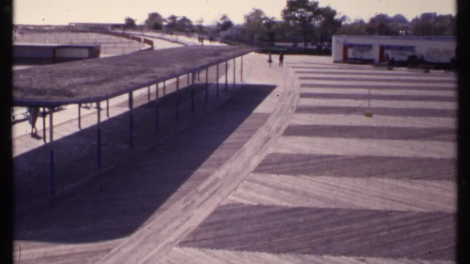 New London 1969 frame showing a beach-adjacent walkway, shaded shelter, low public buildings, trees, and visitors walking in the distance.