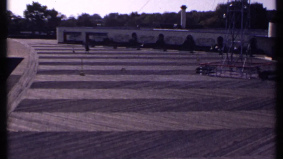 Later 1969 New London frame showing darker exposure over the patterned lot, with distant pedestrians, trees, and a tower beside the low building.