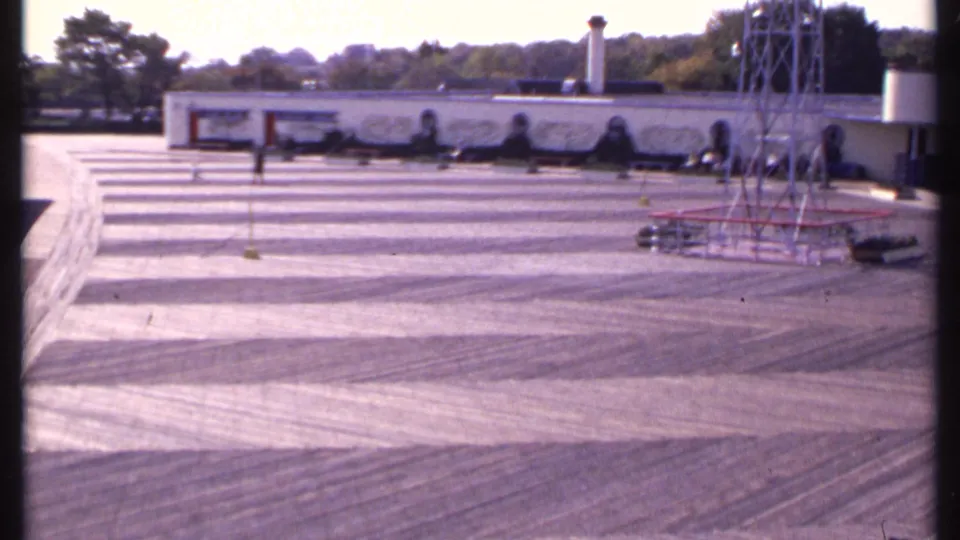 1969 8mm view of a broad New London courtyard with patterned pavement, a low decorated wall, a metal tower, and sparse visitors.