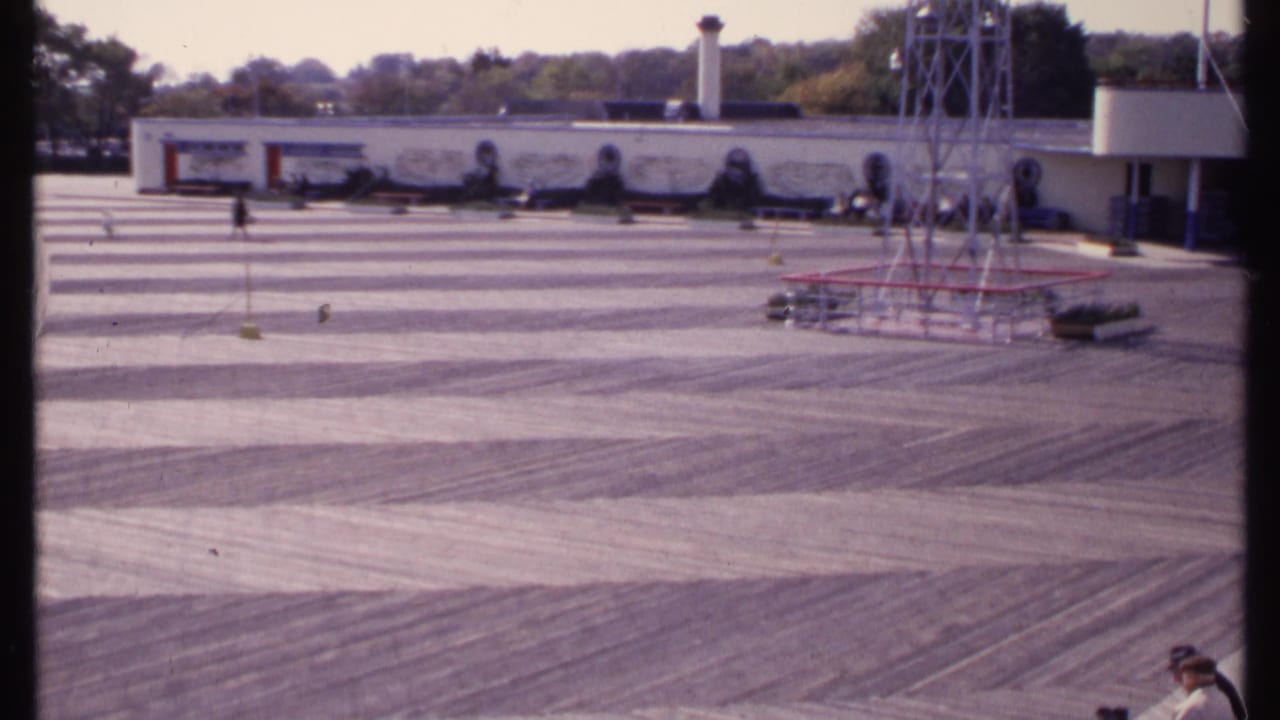 New London courtyard scene on 8mm film, showing a broad paved area, mural-like wall, metal tower, and a person partly visible at right.