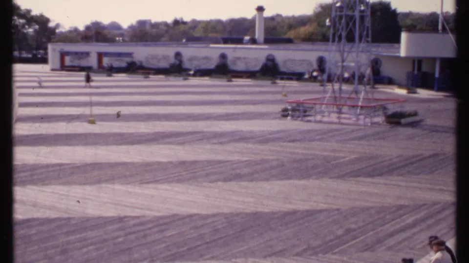 New London courtyard scene on 8mm film, showing a broad paved area, mural-like wall, metal tower, and a person partly visible at right.