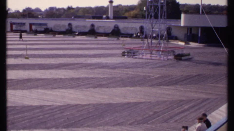 Elevated 1969 home-movie view of the New London courtyard, with patterned paving, a tower frame, mural wall, and seated or standing visitors.