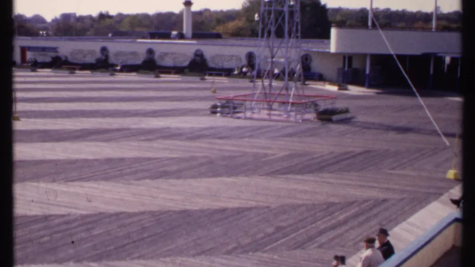 Final 8mm frame of the New London courtyard, showing the tower, mural wall, diagonal line across the view, and visitors near the bottom edge.