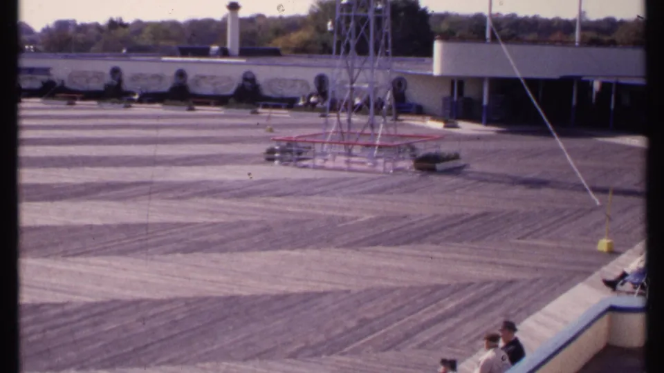 1969 New London home-movie view of a broad outdoor recreation deck with a metal tower, low building, trees, and visitors seated at the edge.