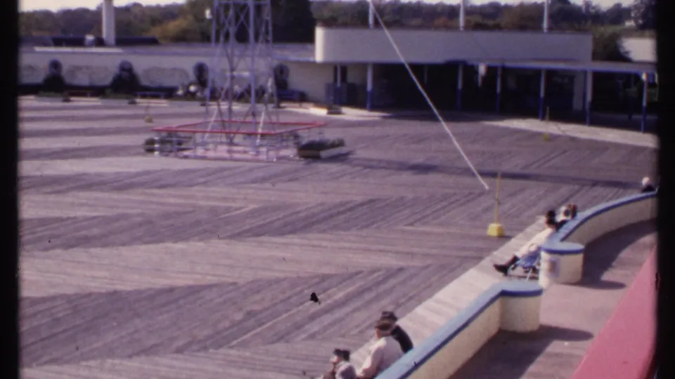1969 New London 8mm frame showing the patterned deck, central metal structure, low building, and visitors seated near the foreground curve.