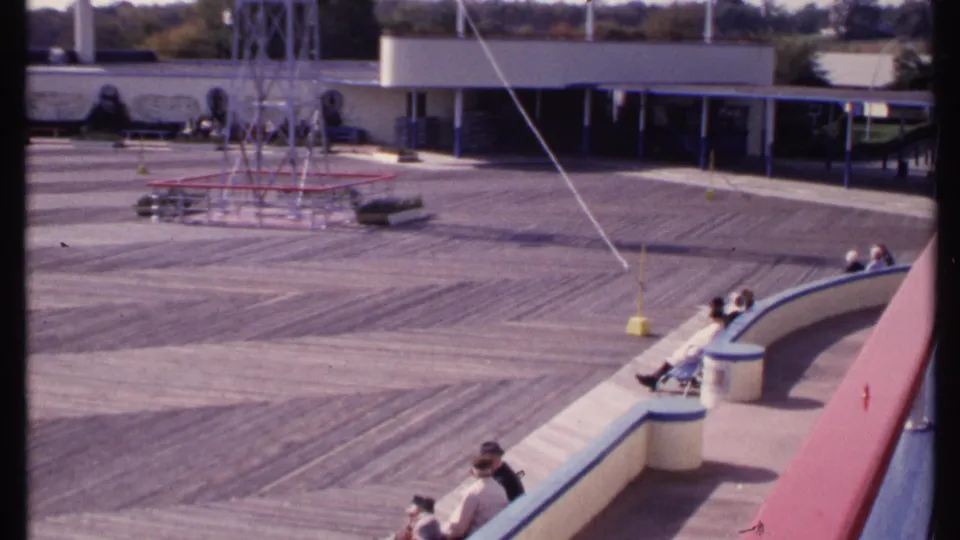 Late-1960s New London home-movie frame of a quiet outdoor recreation deck with visitors seated along a curved bench and a low building behind.