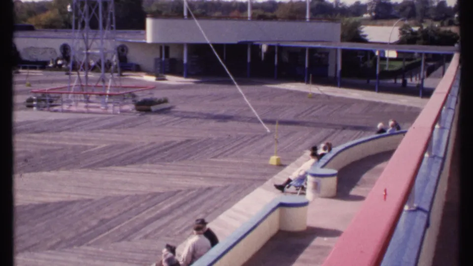 Elevated 1969 New London view of people watching from curved venue seating near a deck, tower, cables, and low building.
