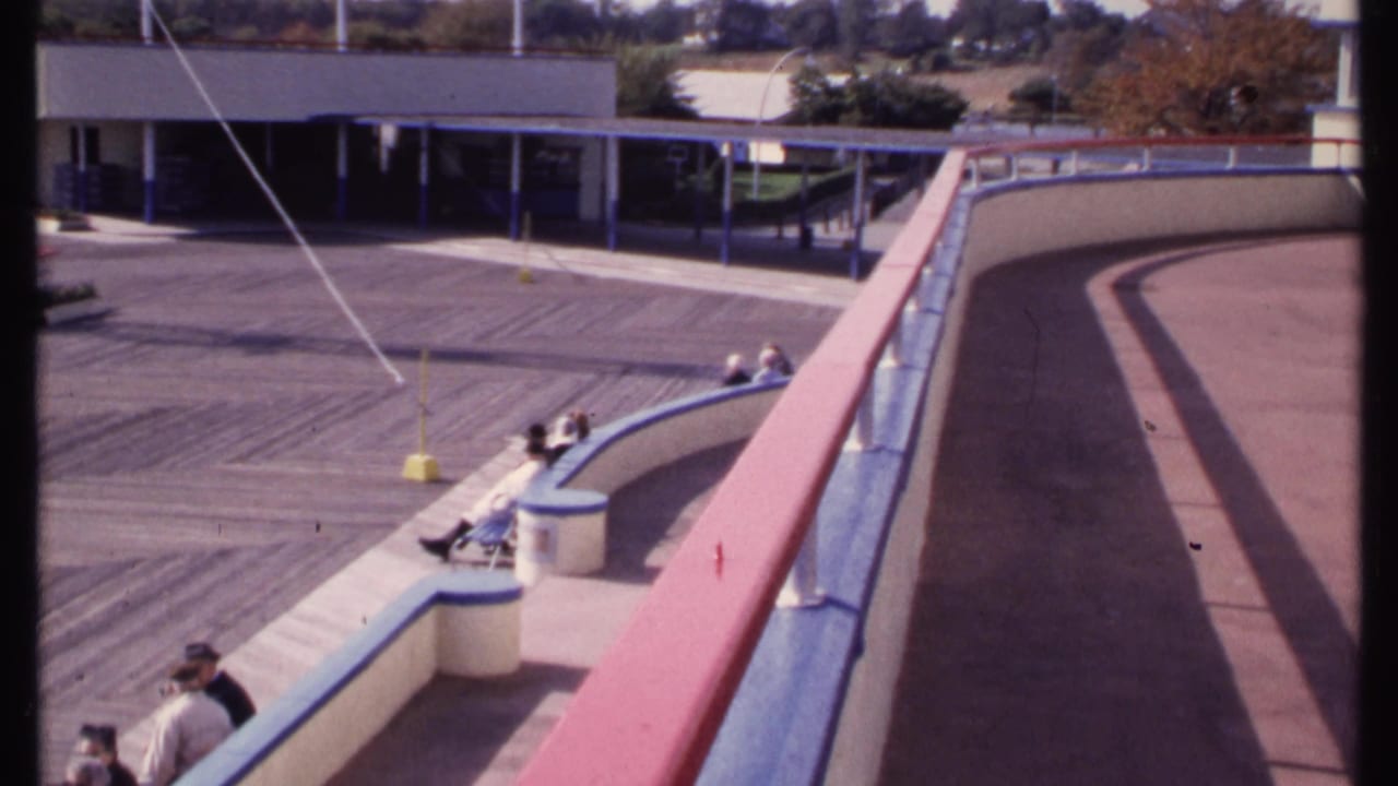 Curved elevated walkway and railing with people seated along the edge near a low building.