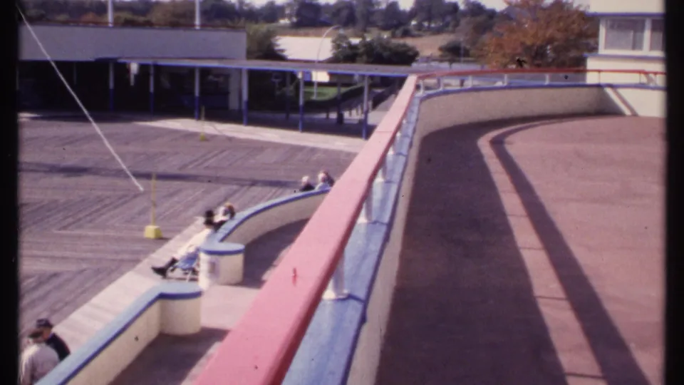 Home-movie frame from New London in 1969 showing a curved boardwalk railing, seated visitors, broad deck surface, and public building beyond.