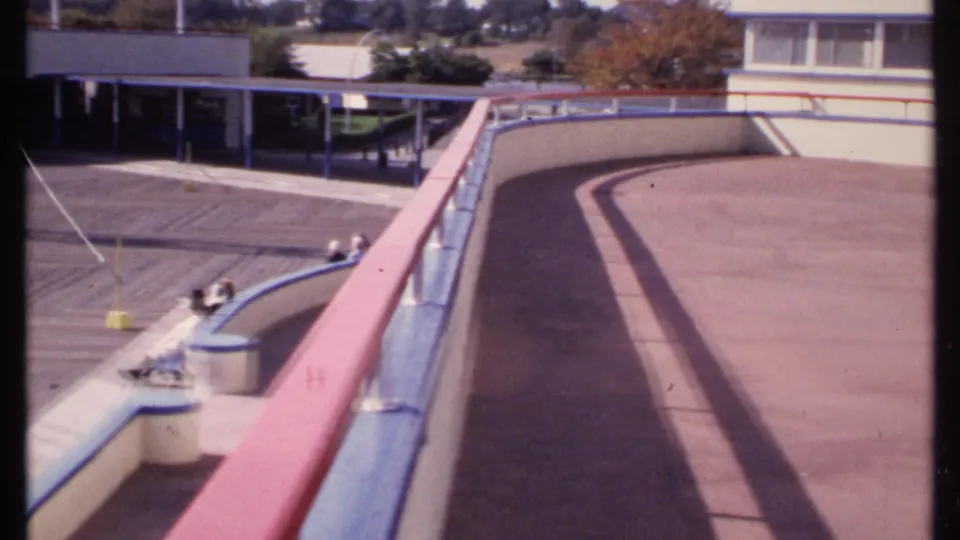 1969 New London boardwalk scene with long railing shadows, seated people in the distance, and a low building across the open paved area.