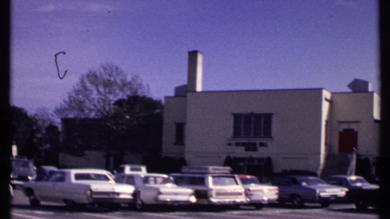 Parking lot with 1960s cars in front of a beige recreation hall building.