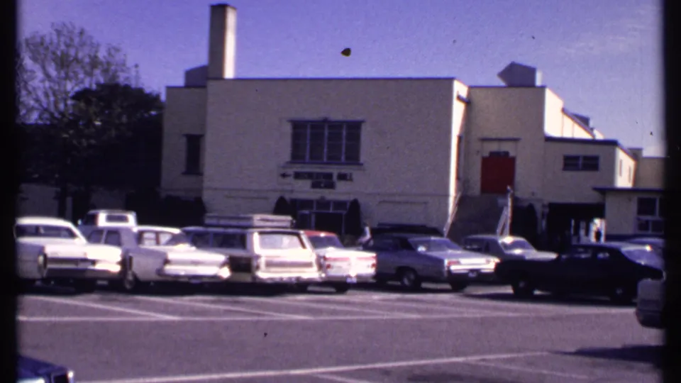 New London 1969 frame with several 1960s cars parked before the beige recreation hall arcade building.