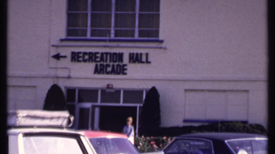 1969 home-movie frame centered on the Recreation Hall Arcade sign, with cars in front and a person at the entrance.