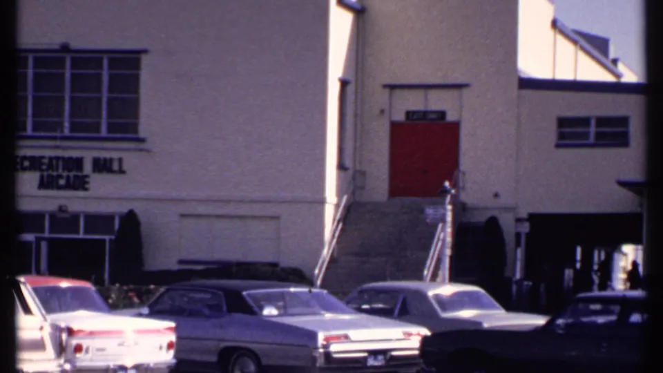 1969 New London home-movie frame showing parked cars in front of a recreation hall or arcade building with red upper doors.