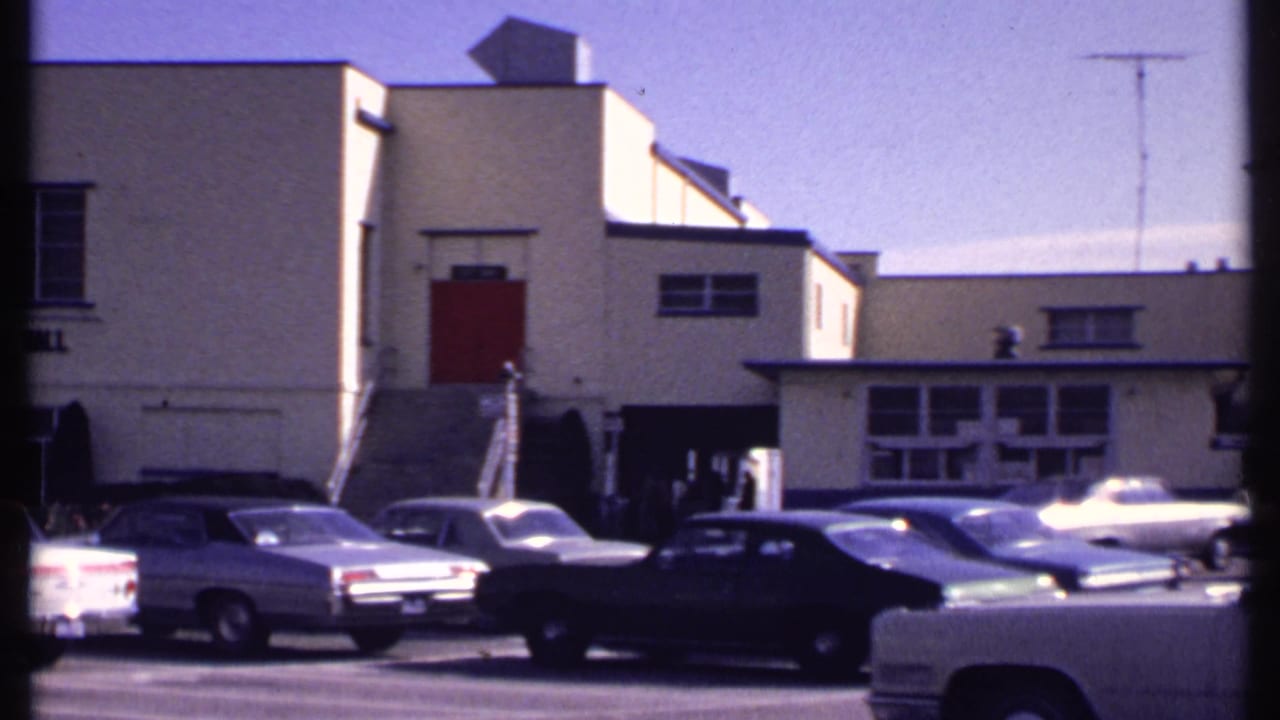 Vintage cars sit in a sunlit New London parking lot as the camera pans past a recreation building with red doors.