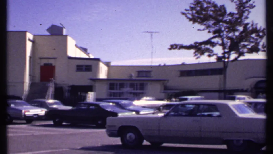 New London 1969 parking lot view with sedans, a station wagon, a low building, and a tree crossing the frame.