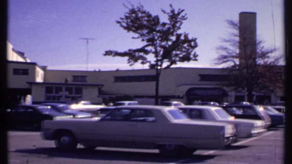 Late-1960s New London frame with a station wagon in the foreground, more parked cars, a tree, and a tall chimney beyond the lot.