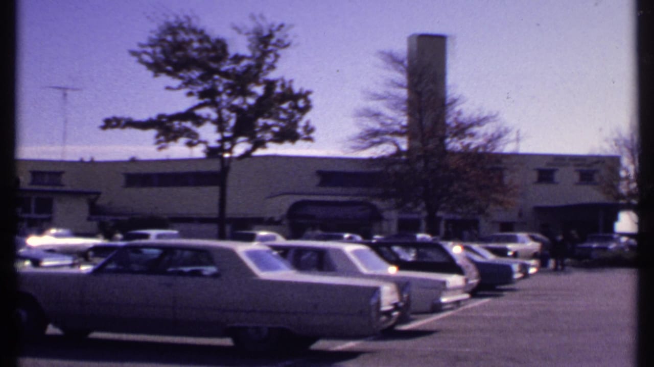 Rows of parked cars face a low building with a tall chimney under a clear sky.