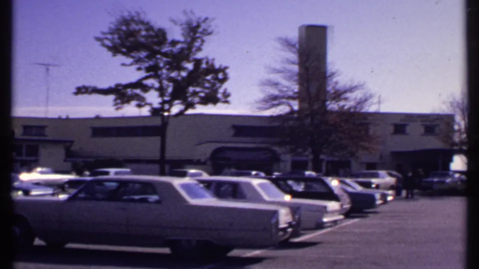 1969 New London parking lot with large American cars, a low commercial building, trees, and a tall chimney in bright daylight.