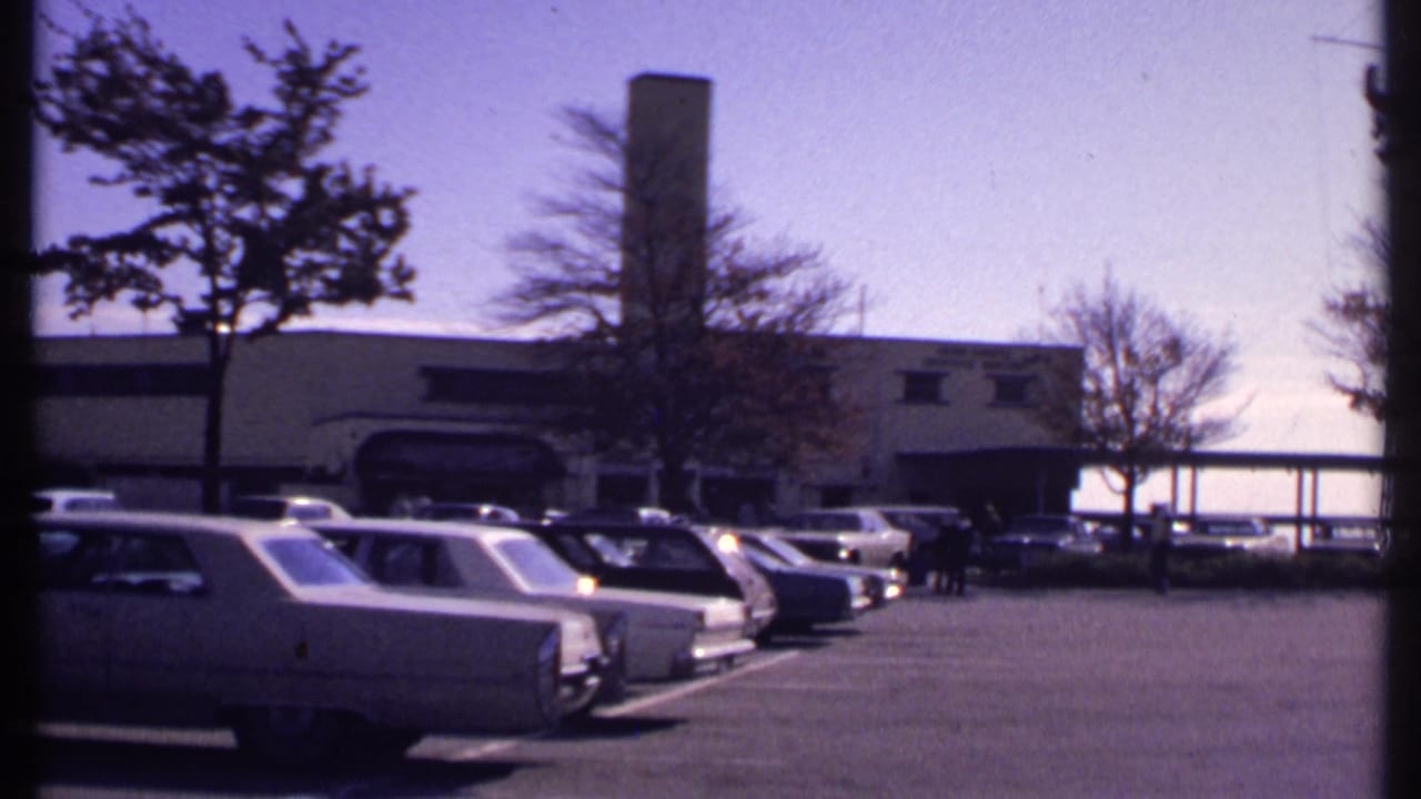 Parked cars sit in rows near a building entrance with a few distant people.