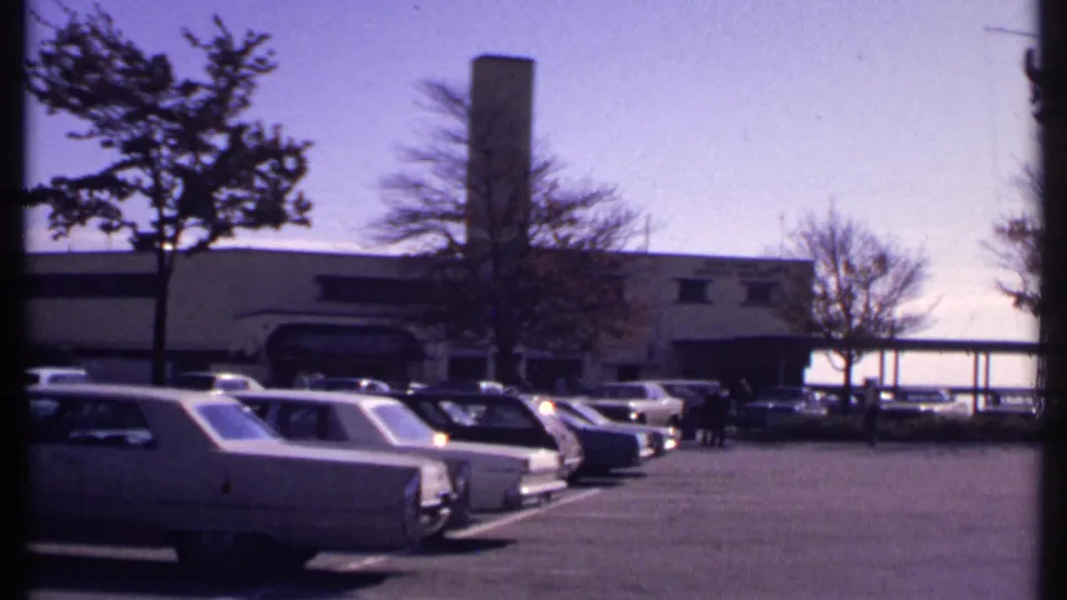 New London, Connecticut parking lot in 1969, showing rows of cars, a low building, trees, and distant pedestrians near the entrance.