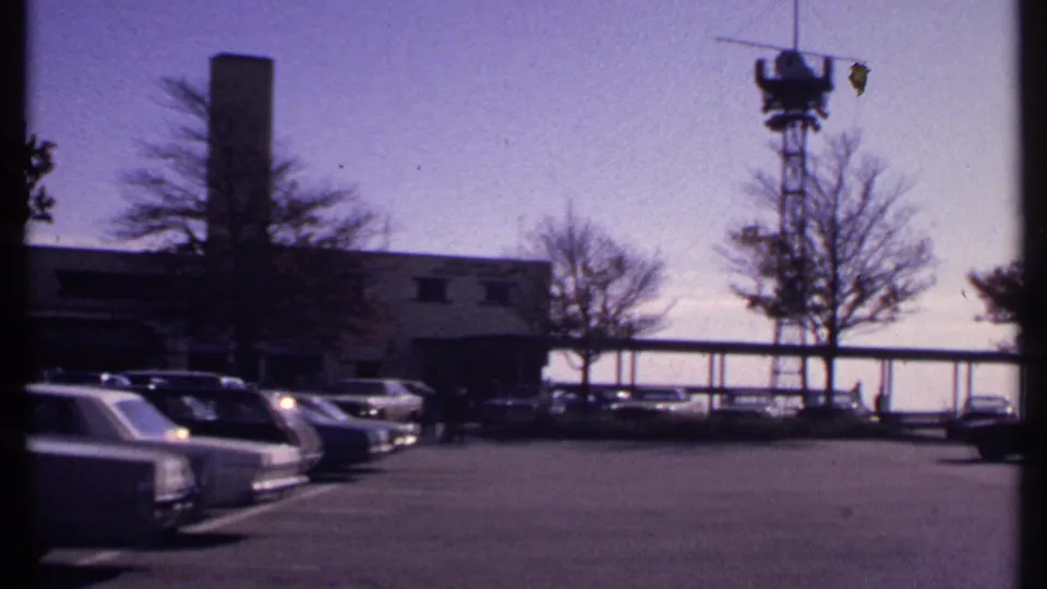 1969 New London parking-lot view with classic cars, a low building, bare trees, and a metal tower visible beyond the pavement.