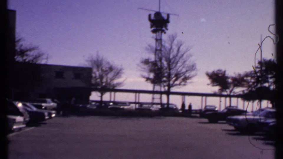 Wide 1969 New London parking-lot frame with cars at the edges, a tower, trees, distant pedestrians, and bright open sky.