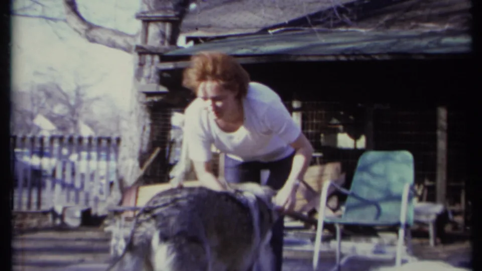 Adult woman in a white shirt bends toward a gray and white dog in a California backyard setting, with a chair and wooden structure nearby.