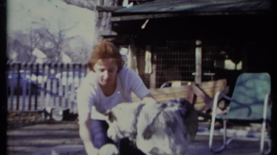 Adult woman crouches close to a gray and white dog in an outdoor California yard, with a green lawn chair and outbuilding behind them.