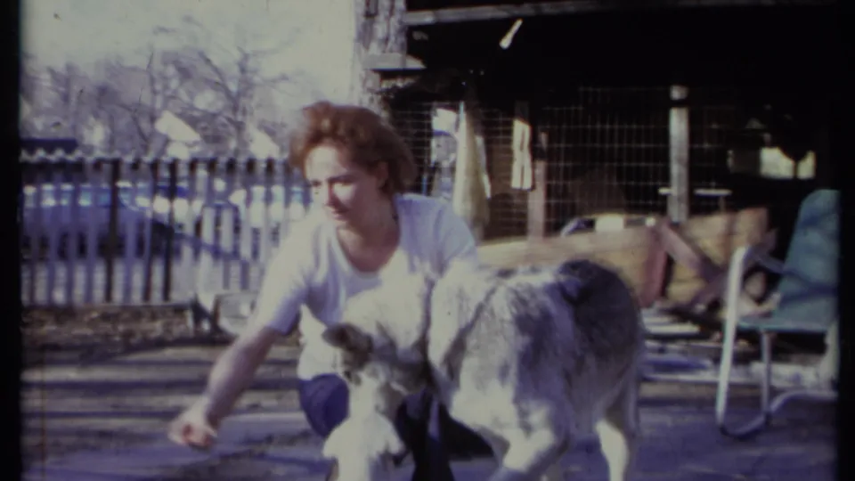 In 1971 California home-movie footage, a woman kneels in a backyard while a gray-and-white dog stands close to her.