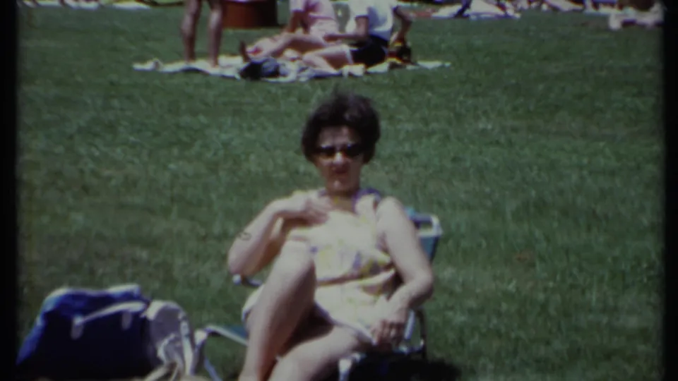In California 1971 home-movie footage, a woman in sunglasses and a yellow patterned dress relaxes in a park chair.
