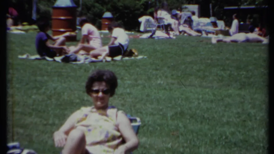 The 8mm frame shows a woman seated in a lawn chair, with casual picnic groups spread across the grass in California.