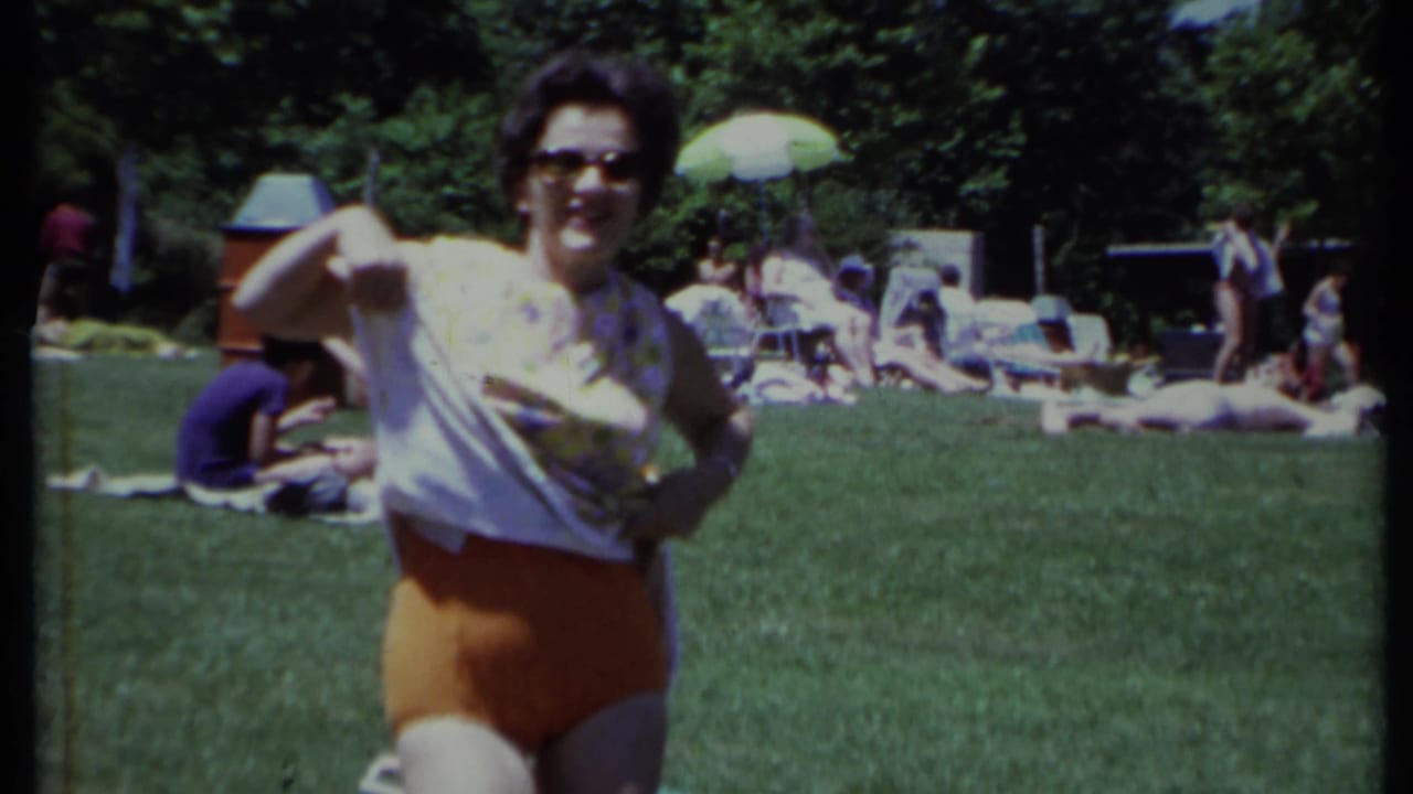 Woman in sunglasses and orange swim bottoms stands on a lawn with people relaxing behind her.