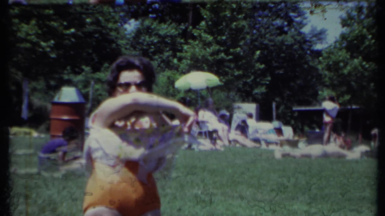 Woman lifts her top while standing on grass, with people and an umbrella in the background.