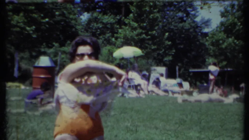 The woman raises her floral top during the outdoor gathering, with sunbathers, trees, and a green umbrella visible behind her.
